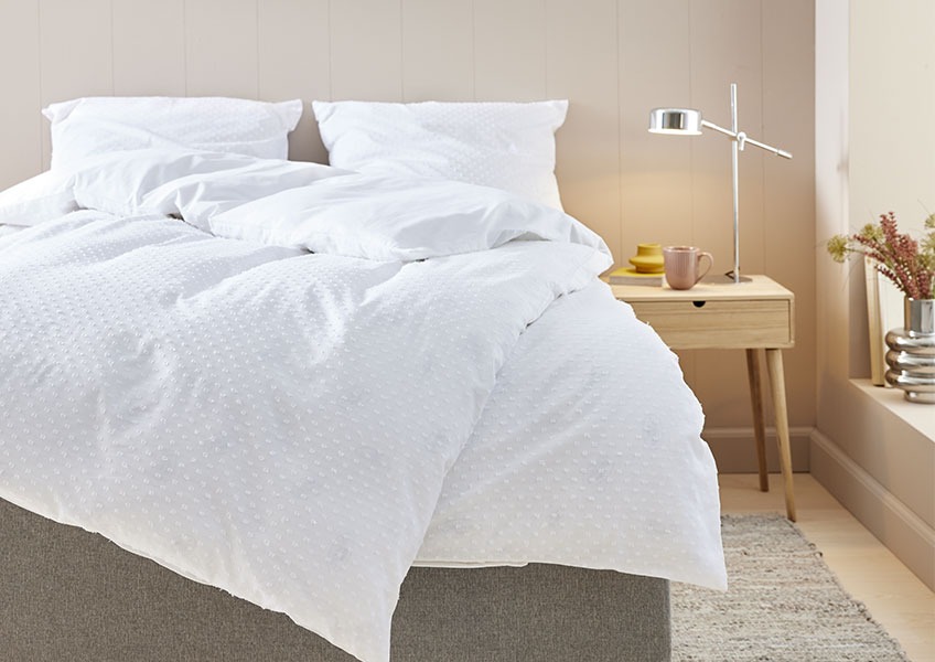 Seng med et hvidt prikket dynebetræk i et soveværelse med sengebord og krom bordlampe