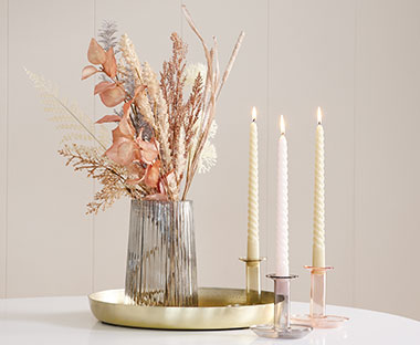 Glasvase med kunstige blomster på gylden pyntebakke og glas lysestager med snoede lys