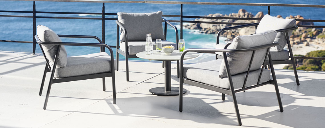 Lounge møbler med stel i aluminium og grå havehynder samt lille rundt bord på solbeskinnet terrasse nær havet