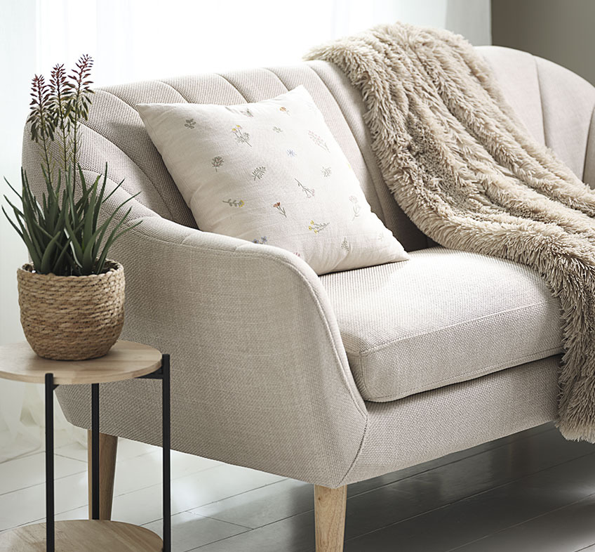 RASMUS kunstig plante i STEFAN blomsterpotte, AGERTIDSEL pyntepude og MYRFIOL plaid Kunstig aloe vera plante med blomster, broderet pyntepude og imiteret pels plaid på beige sofa