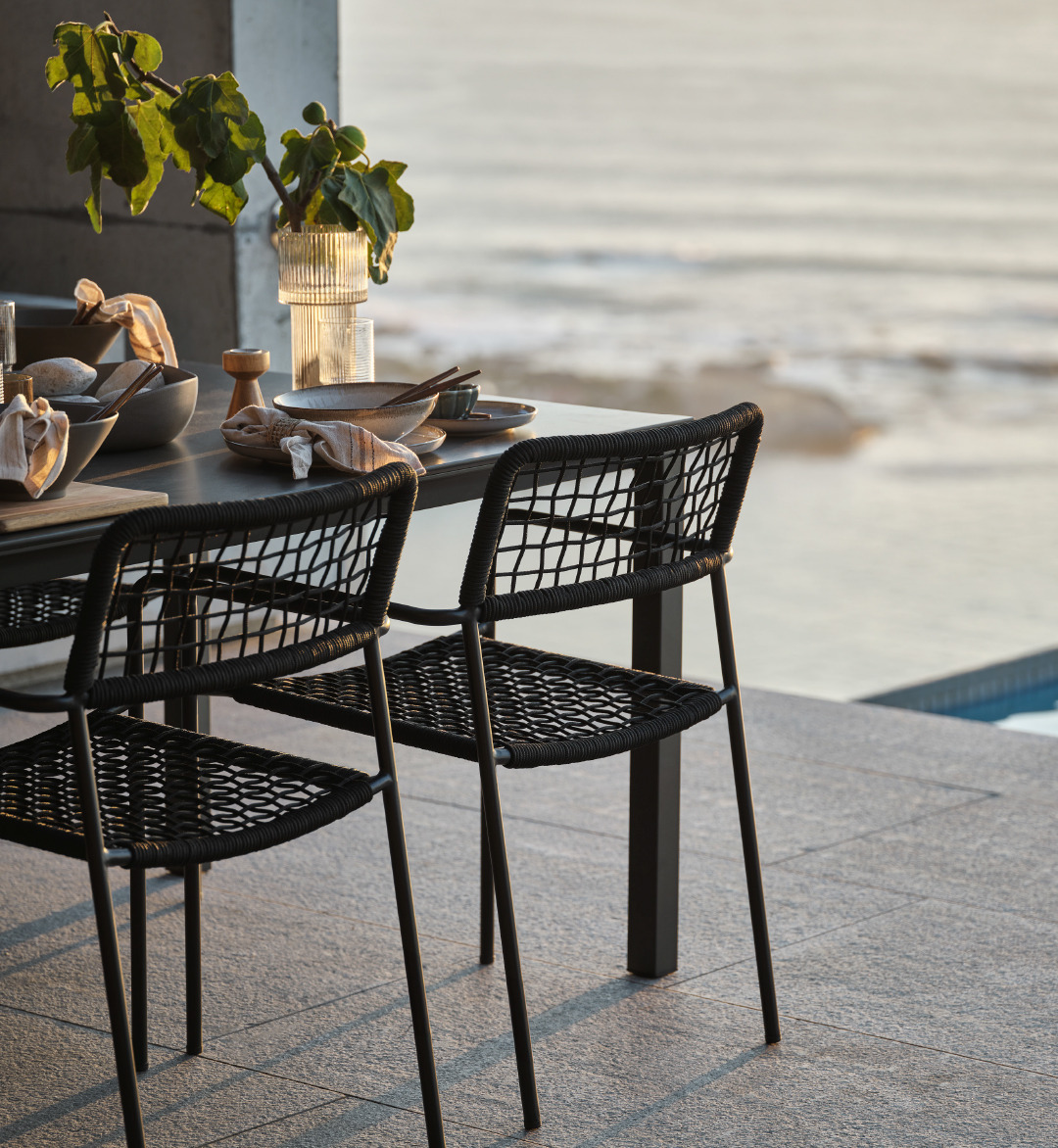 Smukt opdækket havebord LANGET med stabelstol LABING i fokus på terrasse ud til hav og pool