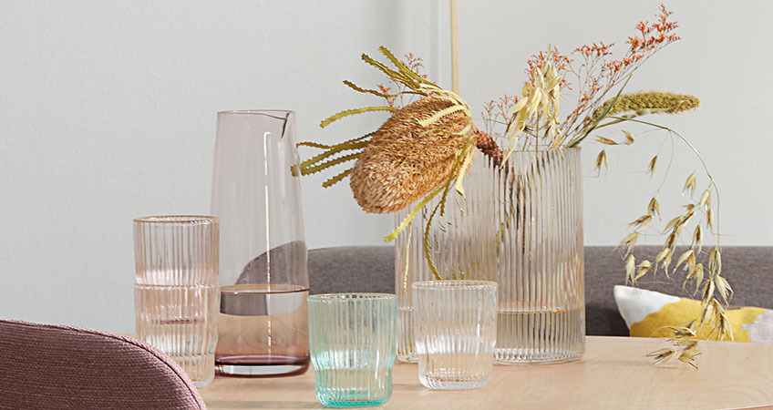 UNO kande, HILMIR vase og CLAES drikkeglas Kande, drikkeglas og vase i farvet glas på et spisebord