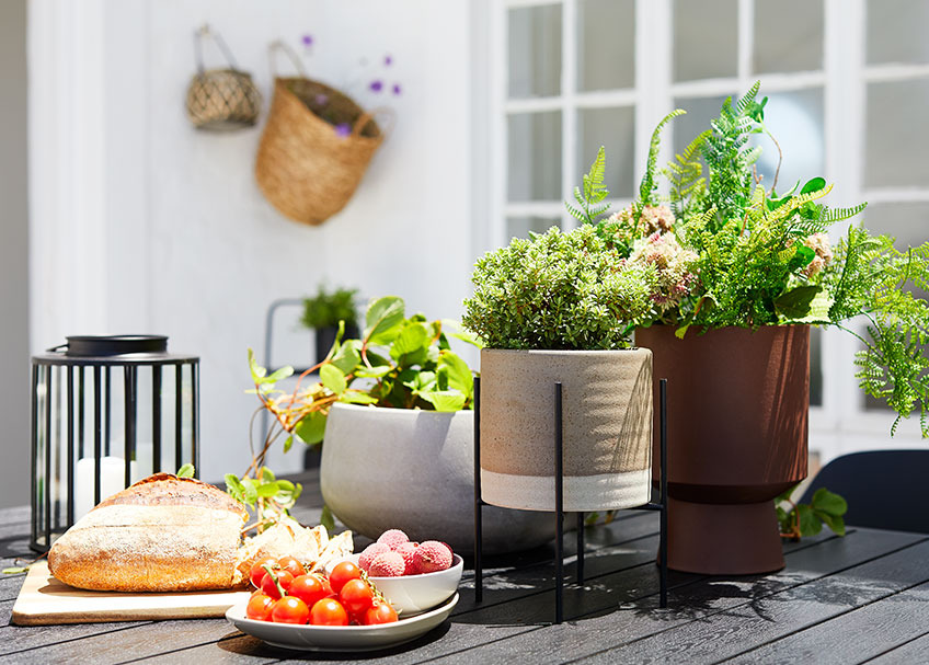 MANDRUP og JOHN blomsterpotter og MYRSANGER lanterne på JERSORE havebord Blomsterpotte i keramik, lanterne og bakke med brød og frugt på et havebord