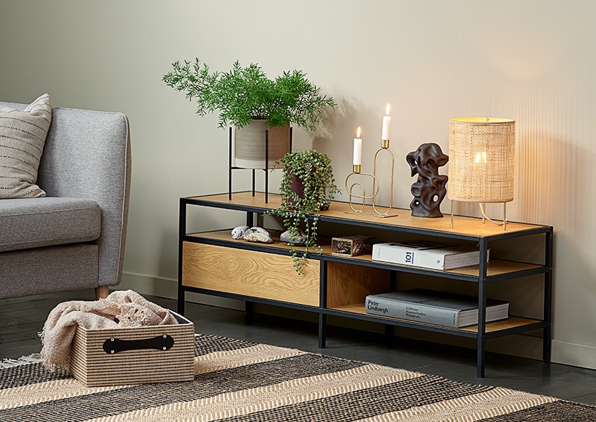 Et hjørne af en stue med en sofa, en kurv, en lille bordlampe og et moderne TV-bord 