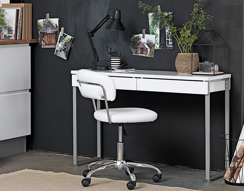 Lille hvidt skrivebord med skuffer og en hvid kontorstol ved en sort væg 
