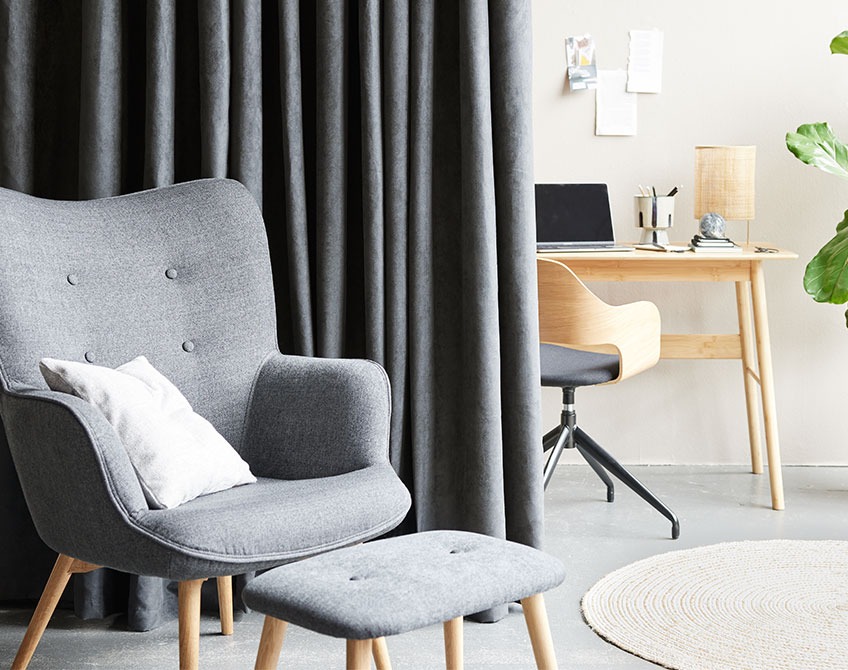Grå gardiner adskiller et hjemmekontor med skrivebord og kontorstol fra en stue med en lænestol og fodskammel