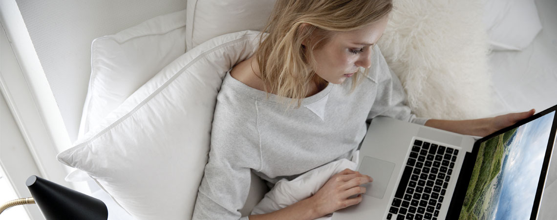 Kvinde sidder med bærbar computer og rygpuder i sengen